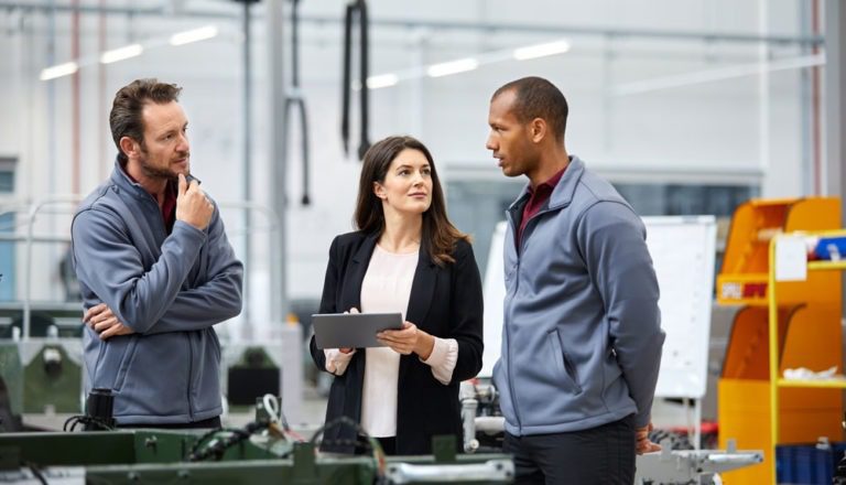 Three professionals standing in a factory and talking, while one woman holds a digital tablet.