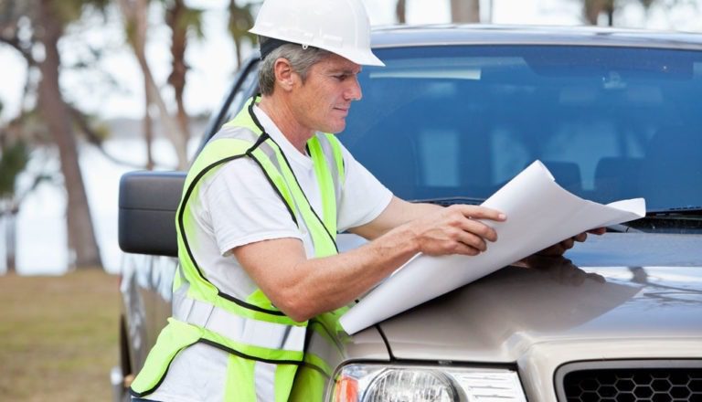 Construction worker in a hard hat looks at building plans on the hood of his pickup truck.