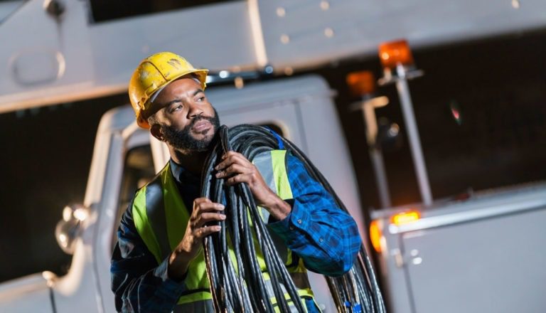 Male technology worker wearing a hard hat and safety vest holding cables outside of a white service truck.