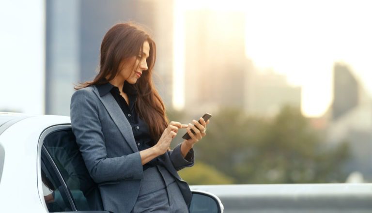 Woman in professional attire using her phone while leaning against her car.